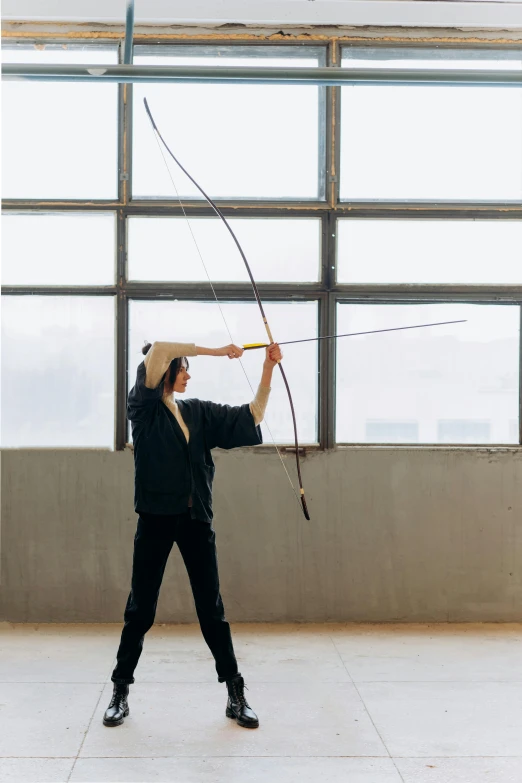 a woman holding a bow and arrow in front of a window, inspired by Fei Danxu, unsplash, arabesque, standing in an arena, helio oiticica, production photo, doing martial arts