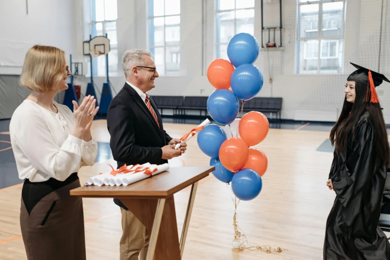 a group of people standing around a table with balloons, pexels contest winner, american barbizon school, orange and blue color scheme, ceremony, amanda lilleston, in the high school gym