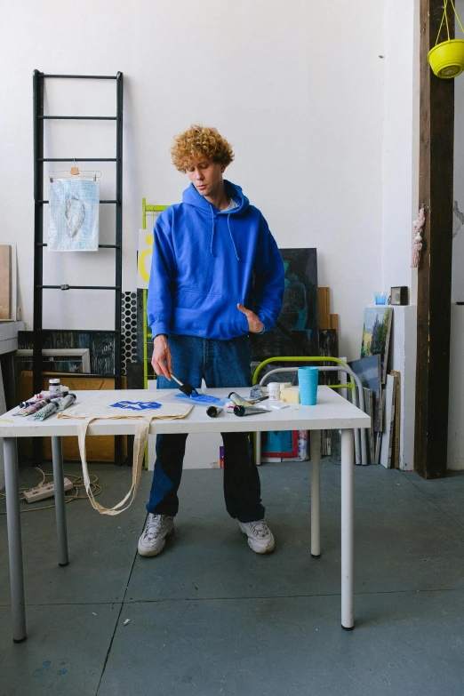 a man standing next to a table in a room, process art, wearing a blue hoodie, commercially ready, teenage boy, dale odell