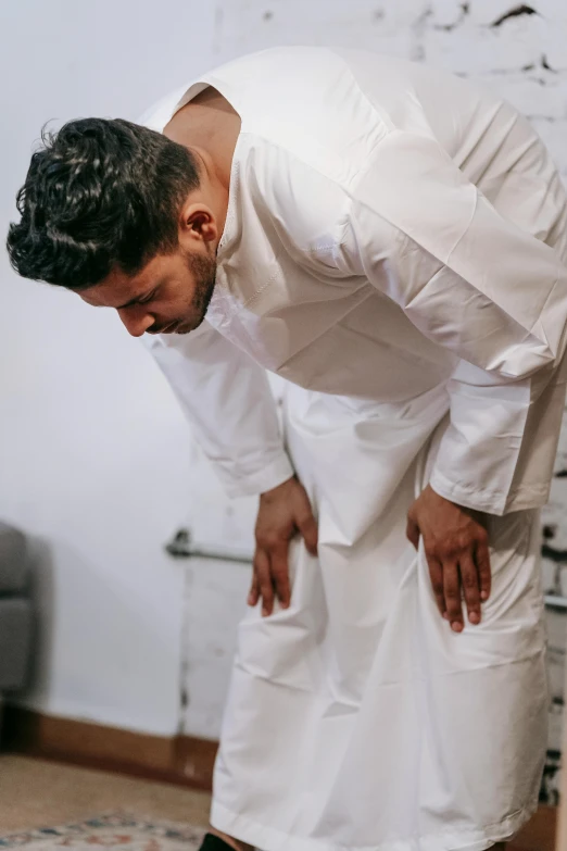 a man standing next to a cat on a rug, wearing a white hospital gown, bend over posture, islamic, lower back