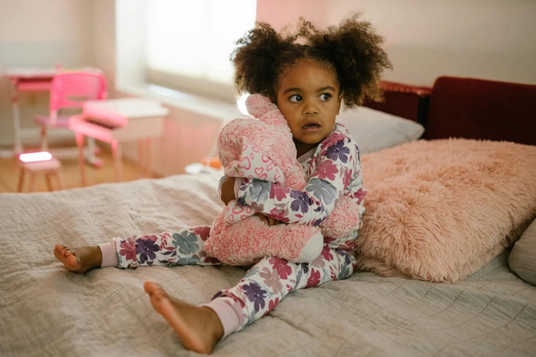 a little girl sitting on top of a bed holding a stuffed animal, inspired by Myles Birket Foster, pexels contest winner, with afro, wearing a tracksuit, pink, patterned