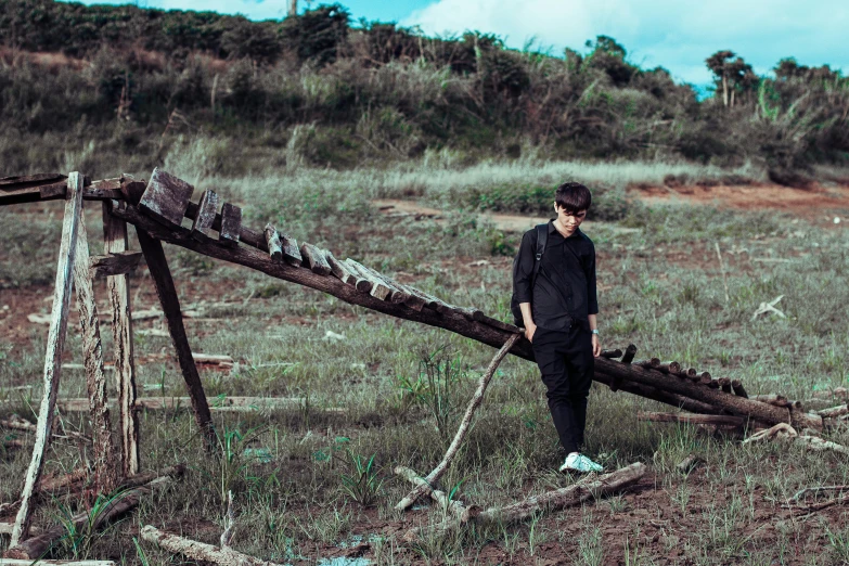 a man standing in a field next to a wooden structure, an album cover, unsplash, conceptual art, standing in a space ship wreck, declan mckenna, rugged black clothes, wooden bridge