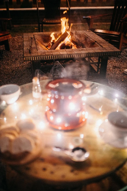 a table topped with bowls of food next to a fire, with a cup of hot chocolate, at night with lights, magic