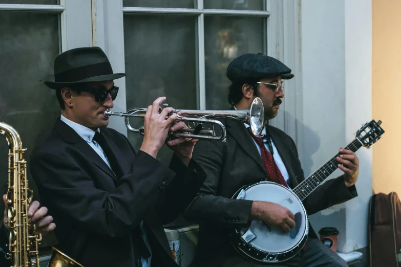 a group of men sitting next to each other playing musical instruments, by Carey Morris, unsplash, wearing victorian brass goggles, pork pie hat, blacks and blues, two men in black