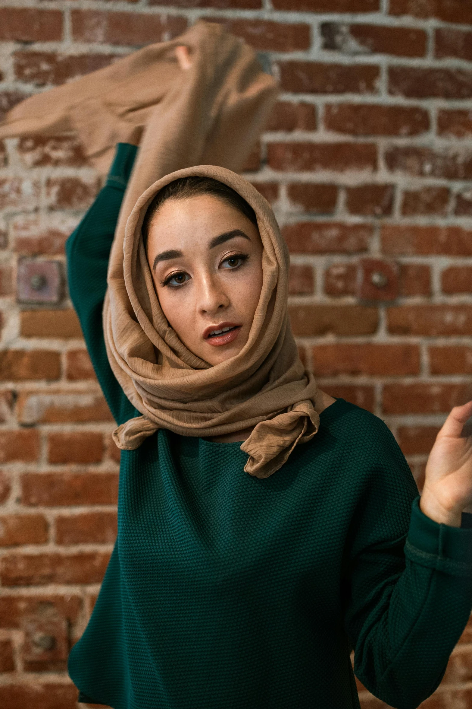 a woman standing in front of a brick wall, inspired by Maryam Hashemi, trending on pexels, hurufiyya, wearing a green sweater, wearing a scarf, light skin tone, expressive pose