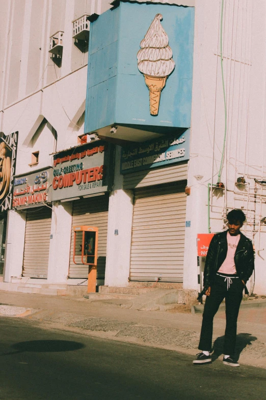 a man standing on the side of a road in front of a building, an album cover, trending on unsplash, ice cream, somalia, hindu aesthetic, 1990's photo
