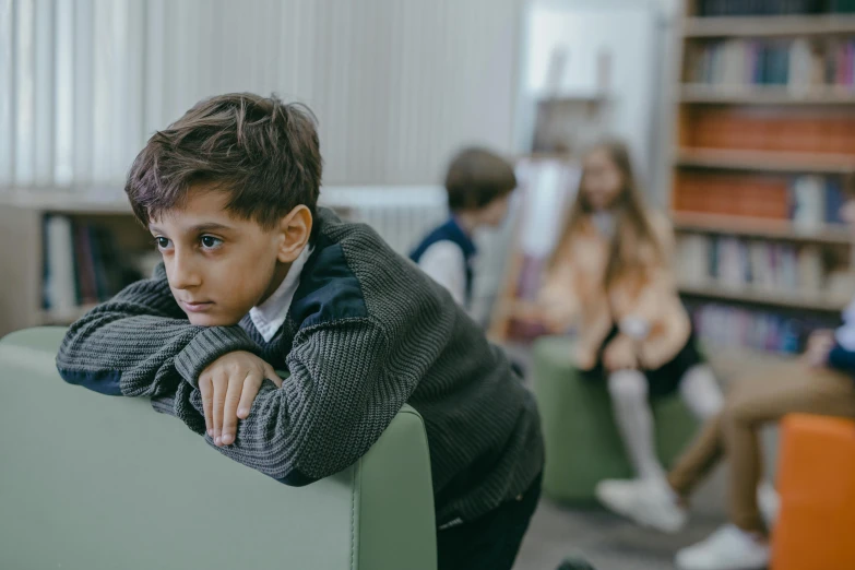 a young boy sitting on a green chair in a library, trending on pexels, incoherents, his friends are angry, in a school classroom, te pae, sitting in a lounge