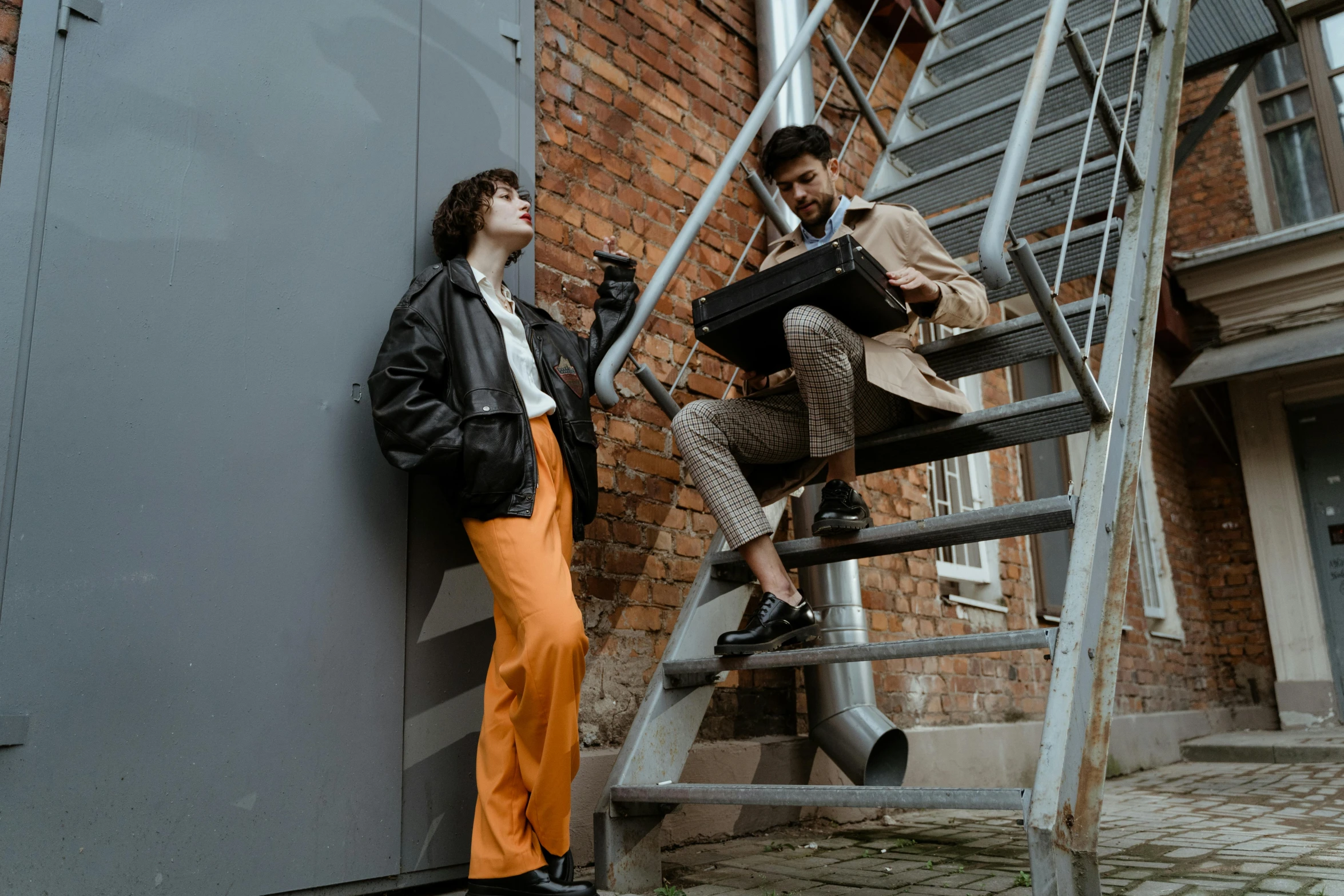 a man and a woman standing on a set of stairs, by Emma Andijewska, trending on pexels, brown pants, industrial colours, thumbnail, black and orange