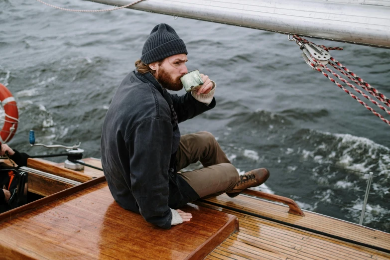 a man sitting on a boat drinking from a cup, he also wears a grey beanie, he's on an old sailing boat, cold brew coffee ), lachlan bailey