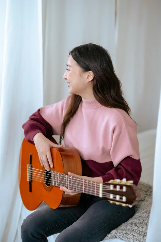 a woman sitting on a bed playing a guitar, inspired by Song Xu, pexels contest winner, happening, wearing casual sweater, brown and pink color scheme, profile pic, young asian woman