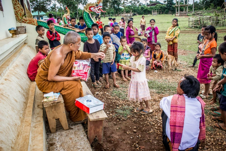 a monk sitting on a bench in front of a group of children, by Daniel Lieske, pexels contest winner, happening, giving gifts to people, thawan duchanee, square, ground breaking