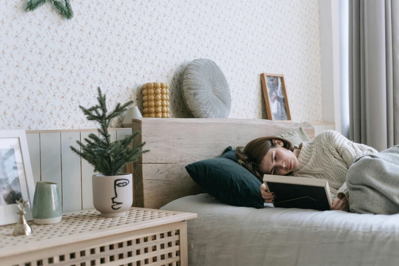 a woman laying on a bed reading a book, a cartoon, inspired by Elsa Bleda, trending on pexels, small and cosy student bedroom, looking tired, a wooden, bedhead