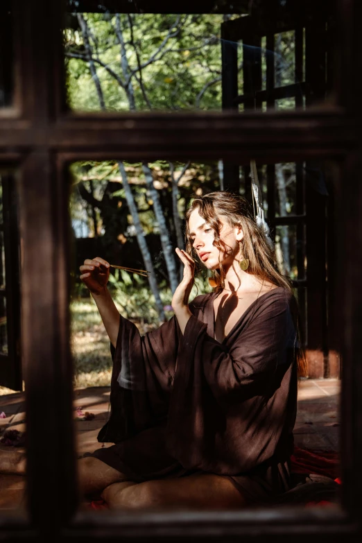 a woman sitting in front of a window talking on a cell phone, a picture, by Julia Pishtar, pexels contest winner, beautiful wood elf, brown robes, in the yard, looking in mirror