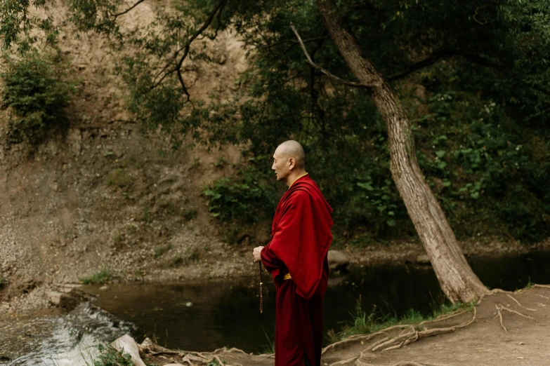a man in a red robe standing next to a river, a portrait, unsplash, process art, monk meditate, next to a tree, profile image, wearing mage robes