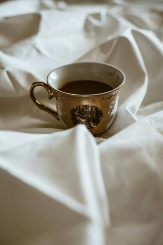 a cup of coffee sitting on top of a bed, gold silk, wrinkled, on a white table, old english