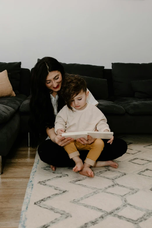 a woman and child sitting on a rug in a living room, holding a book, slightly minimal, riyahd cassiem, jen atkin