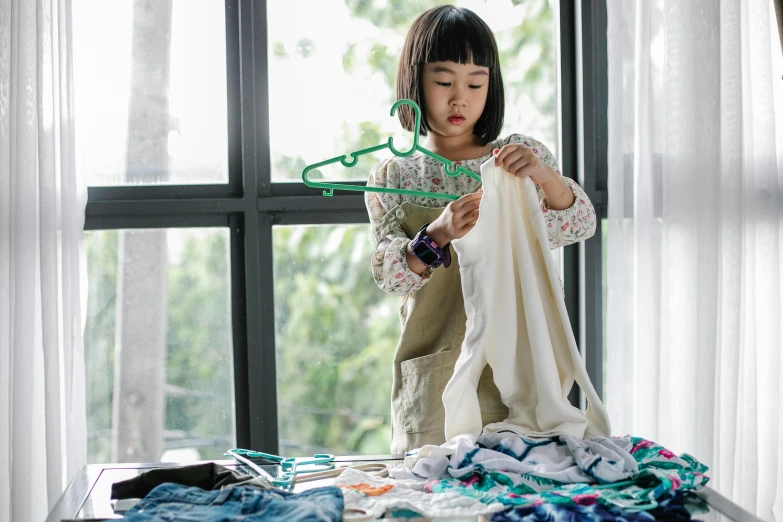 a little girl that is standing in front of a window, laundry hanging, sea - green and white clothes, asian descent, working hard