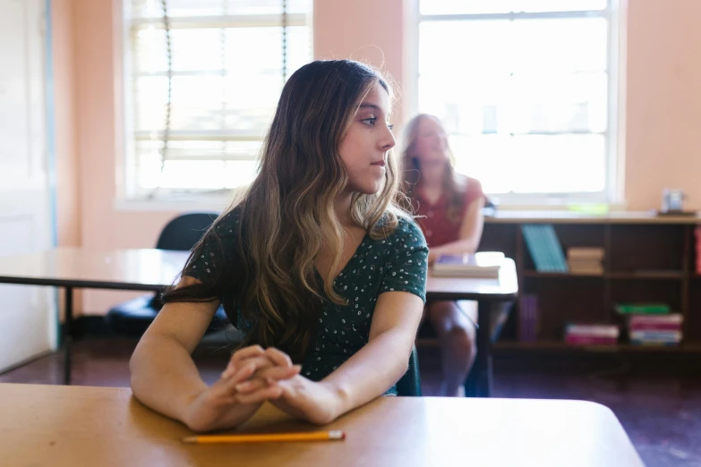 a woman sitting at a table in a classroom, by Anna Findlay, pexels, with a figure in the background, teen girl, background image, pensive