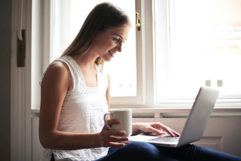 a woman sitting in front of a window using a laptop, pexels, with a white mug, handsome girl, with intricate detail, official screenshot