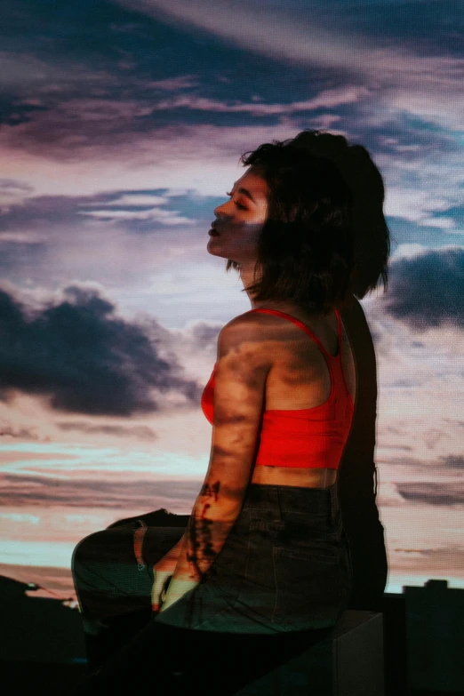 a woman sitting on a ledge with a sunset in the background, an album cover, trending on pexels, art photography, tattooed, charli xcx, on clouds, profile image