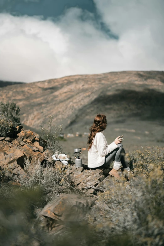 a woman sitting on top of a rocky hill, by Sara Saftleven, trending on unsplash, tea, white, grey, outdoors mesa setting