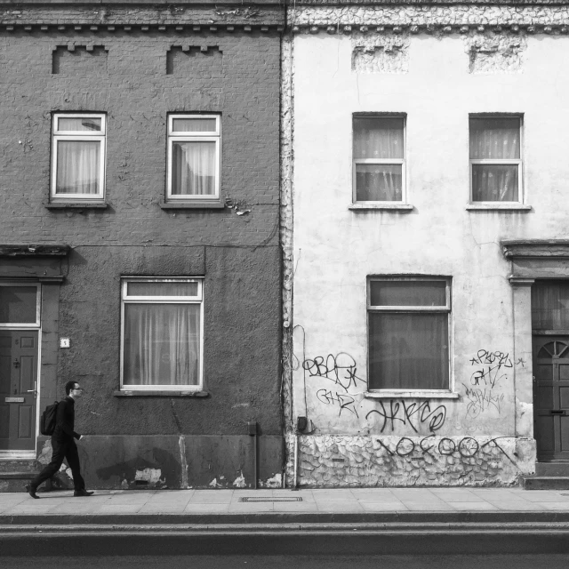 a black and white photo of a man walking down the street, by Kevin Connor, pexels contest winner, postminimalism, dilapidated houses, then another, patrick o'keeffe, two tone