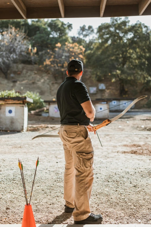 a man in a black shirt and khaki pants holding a pair of arrows, softair arena landscape, camp, bows, top