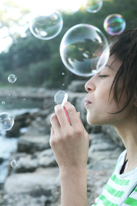 a woman blowing bubbles in front of a body of water, a young asian woman, profile image, small, large)}]