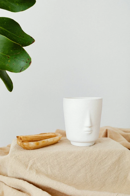 a white cup sitting on top of a table next to a plant, an abstract sculpture, smooth facial features, product photo, linen, drinking