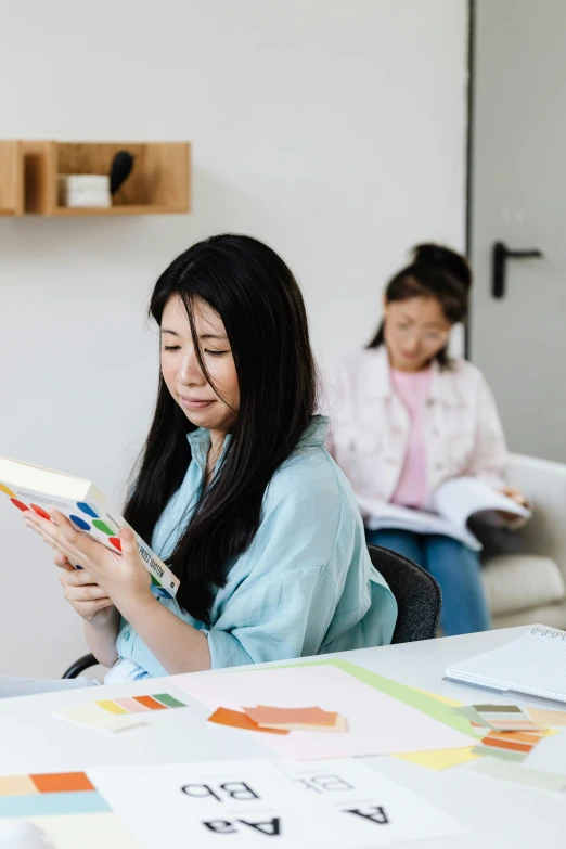 a woman sitting at a table reading a book, a cartoon, by Jang Seung-eop, pexels contest winner, in the background, educational supplies, two buddies sitting in a room, asian girl with long hair