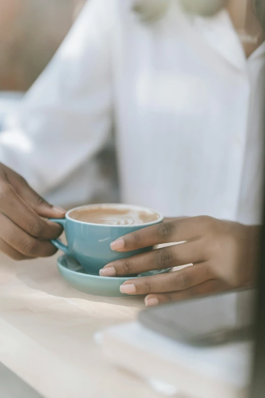 a woman sitting at a table with a cup of coffee, teal, holding hands, wearing a light blue shirt, soft light from the side