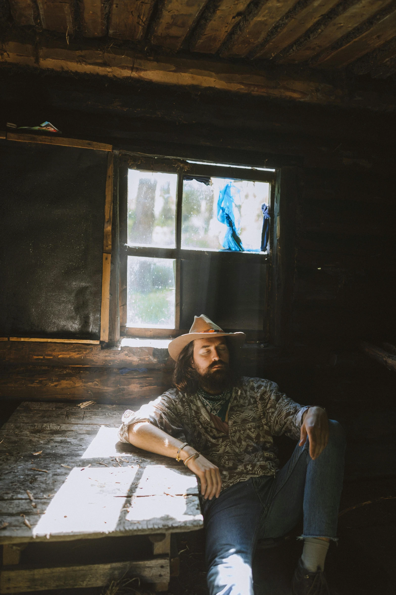 a man sitting at a table in front of a window, a portrait, by Winona Nelson, unsplash, bearded cowboy, album cover, kneeling at the shiny floor, cabin