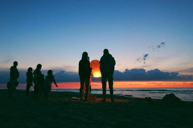 a group of people standing on top of a sandy beach, holding a lantern, profile image, families playing, fire on the horizon