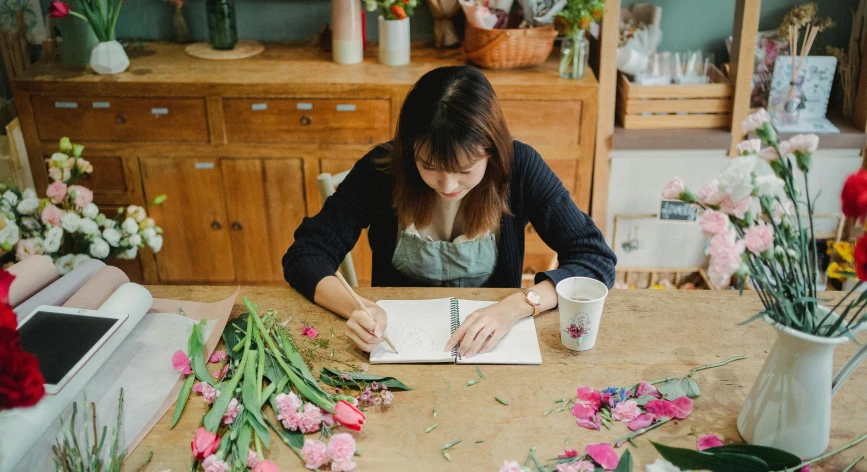 a woman sitting at a table writing on a piece of paper, a still life, by Alice Mason, pexels contest winner, japanese flower arrangements, in a workshop, thumbnail, 1614572159