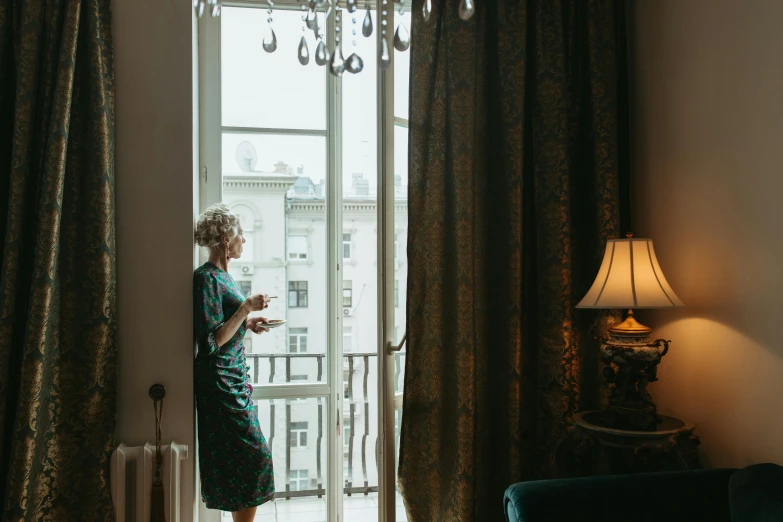 a woman in a green dress standing in front of a window, by Emma Andijewska, pexels contest winner, art nouveau, hotel room, morning coffee, moody : : wes anderson, a blond