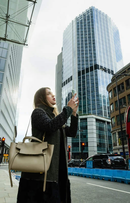 a woman standing on a city street taking a picture, with tall glass skyscrapers, in london, f/1.4, square