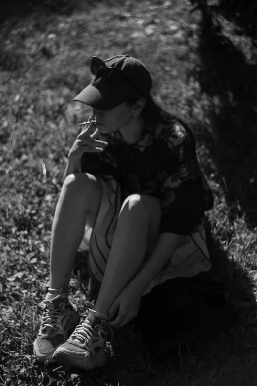 a black and white photo of a person sitting in the grass, tumblr, realism, smoking woman, baggy clothing and hat, in style of petra collins, yung lean