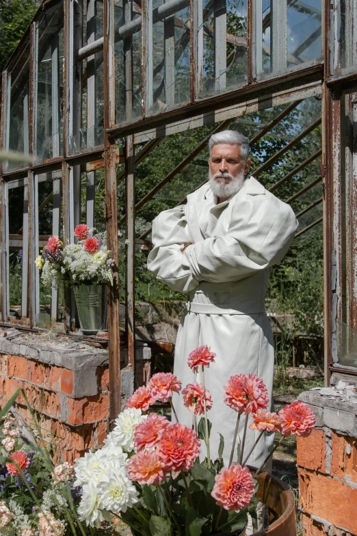 a man standing in front of a window next to flowers, inspired by Illarion Pryanishnikov, wearing long silver robes, in bloom greenhouse, overalls and a white beard, press shot