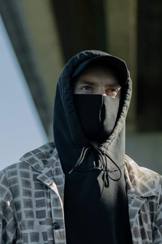 a man wearing a face mask standing in front of a bridge, an album cover, by Joseph Severn, hooded cowl, detail shot, black clothing, caucasian