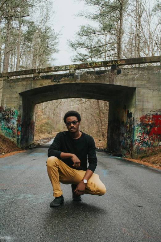 a man squatting in the middle of a road, an album cover, inspired by Gordon Parks, graffiti, in style of tyler mitchell, on a bridge, standing on a forest, j. h. williams iii