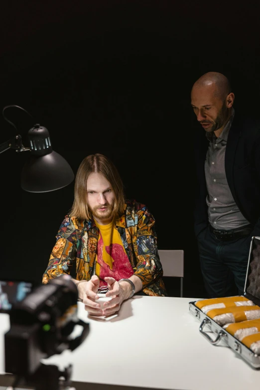 a man and a woman sitting at a table looking at a cell phone, a hyperrealistic painting, inspired by Daniël Mijtens, unsplash, hyperrealism, in a dark studio room, wes anderson and gucci, panavision, behind the scenes photo