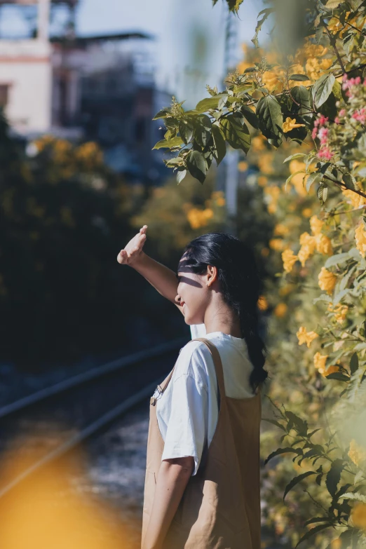 a woman standing on the side of a train track, unsplash contest winner, with yellow flowers around it, greeting hand on head, with soft bushes, vietnamese woman