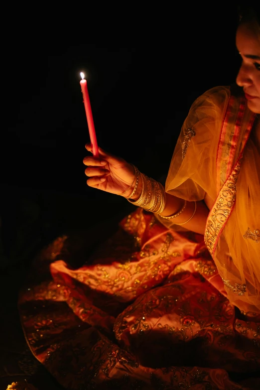 a woman sitting on the ground holding a lit candle, dressed in a sari, lights off, promo image, slide show