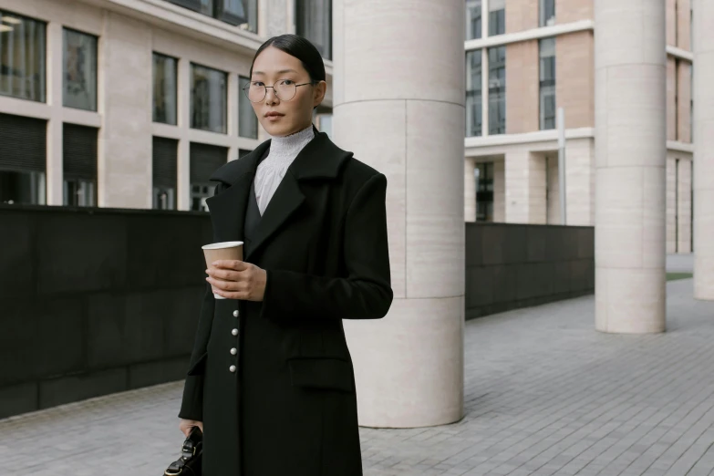 a woman standing on a sidewalk holding a cup of coffee, inspired by Fei Danxu, pexels contest winner, wearing a black noble suit, wearing headmistress uniform, wearing a turtleneck and jacket, architect