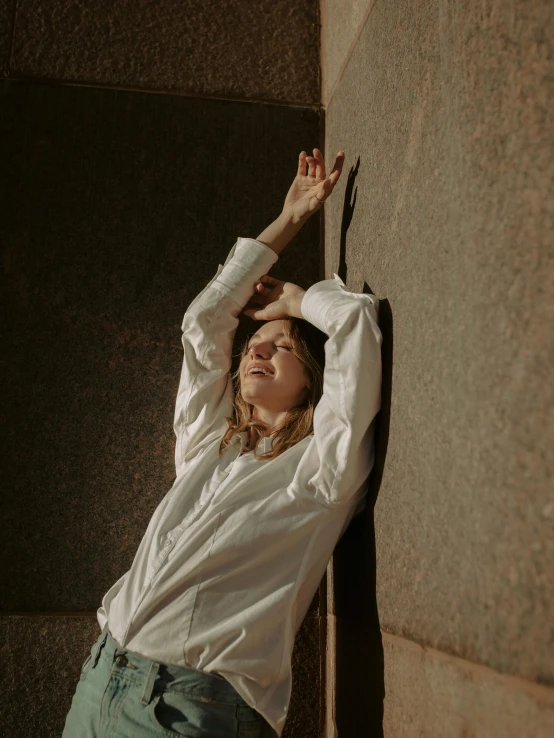 a woman leaning against a wall with her hands on her head, inspired by Elsa Bleda, unsplash contest winner, happening, wearing a white button up shirt, lie on a golden stone, triumphant pose, long sleeves