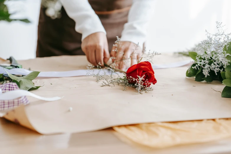 a woman is arranging flowers on a table, by Julia Pishtar, pexels contest winner, holding a bow, on parchment, red rose, sustainable materials