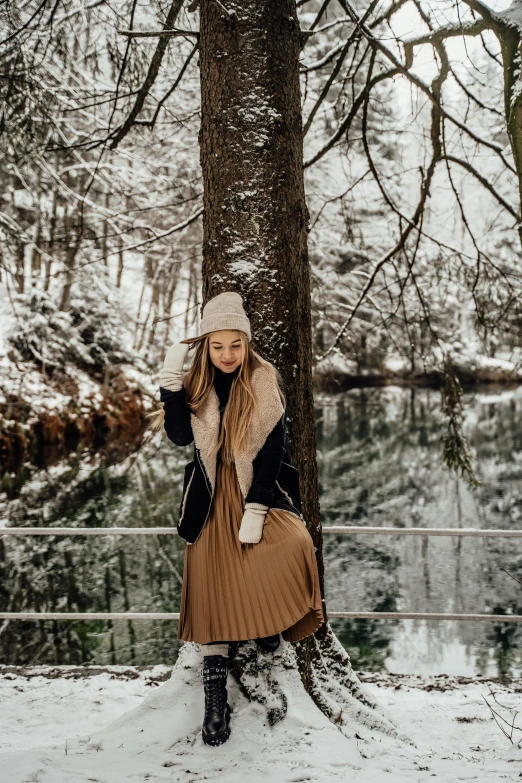a woman standing next to a tree in the snow, pexels contest winner, pleated skirt, brown and cream color scheme, tourist photo, wearing beanie
