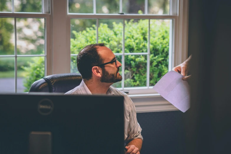 a man sitting at a desk in front of a computer, by Carey Morris, pexels contest winner, in meeting together, looking around a corner, lachlan bailey, next to window