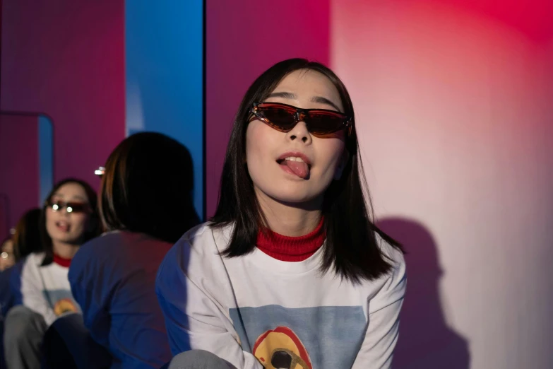 a woman sitting in front of a mirror wearing sunglasses, an album cover, inspired by Russell Dongjun Lu, trending on pexels, aestheticism, red and blue neon, asian girl, graphic tees, performing a music video