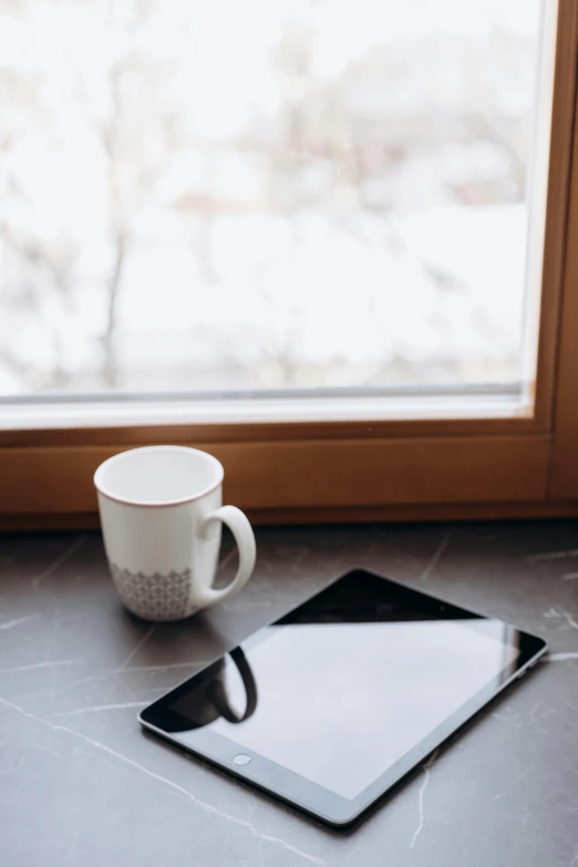 a tablet computer sitting on top of a table next to a cup of coffee, trending on pexels, sitting on a window sill, winter, kitchen counter, ceramics